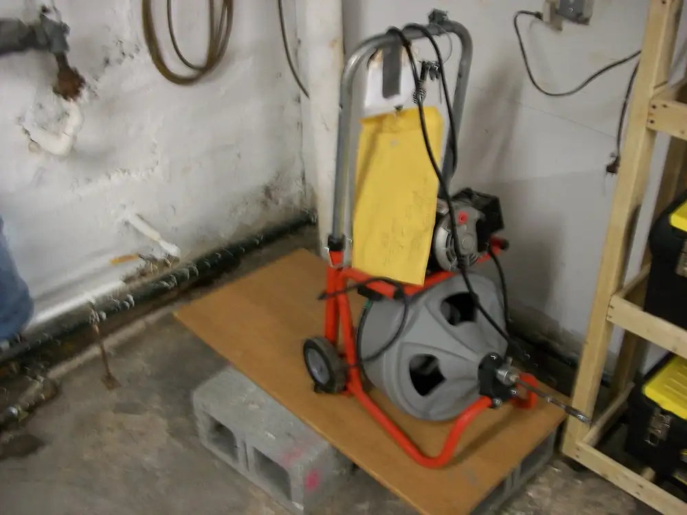 Máquina desatascadora eléctrica de cable, con bobina y ruedas, en un sótano junto a tuberías de desagüe